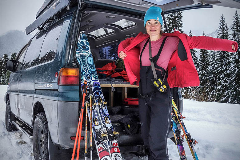 A women pulls on a jacket over her ski bibs and avalanche beacon that's strapped around her torso